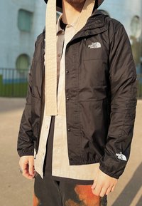 Man som bär en lager på lager-beige skjorta, en svart North Face vindjacka och batikmönstrade byxor, stående utomhus i solljus.