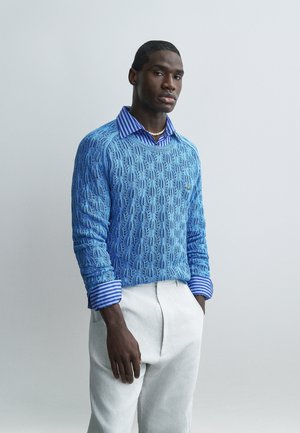 Άνδρας που φοράει μπλε σχέδιο πουλόβερ πάνω από μπλε ριγέ πουκάμισο και λευκό παντελόνι, στέκεται μπροστά σε έναν απλό φωτεινό τοίχο.