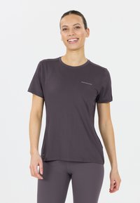 Hymyilevä nainen, jolla on yllään hyvin istuva vaalean violetin värinen lyhythihainen urheilupaita ja siihen matchaavat leggingsit, seisoo valkoista taustaa vasten.