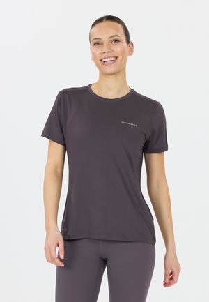 Femme souriante portant un haut de sport ajusté à manches courtes violet et un legging assorti, se tenant devant un fond blanc.