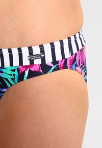 Bikini-Hose mit einem mehrfarbigen tropischen Muster auf schwarzem Hintergrund, kontrastiert durch einen gestreiften Bund. Enthält ein silbernes Logodetail.