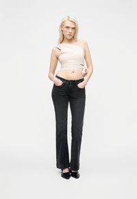 Νεαρή γυναίκα που φοράει μπεζ crop top με έναν ώμο, μαύρο τζιν και μαύρα μυτερά παπούτσια, στέκεται με τα χέρια στις τσέπες μπροστά σε λευκό φόντο.