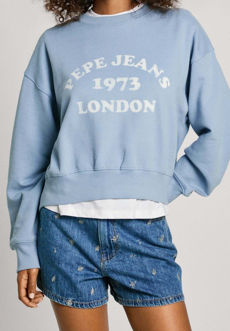 Personne portant un sweat-shirt bleu clair avec le texte "PEPE JEANS 1973 LONDON" et un short en jean bleu avec de petits détails brodés.