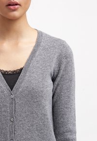 Grå cardigan med V-ringning, enkla knappar och ribbade ärmslut. Mjuk, stickad textur och lager på ett svart topp med spetsdetaljer.