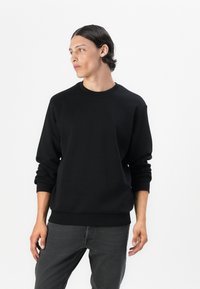Black crewneck sweatshirt in cotton blend with ribbed cuffs and hem, featuring a relaxed fit and smooth texture. Model wears grey jeans.