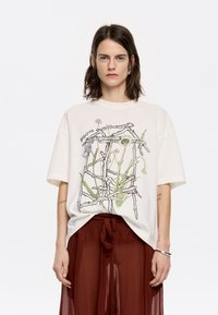 Cremefarbenes T-Shirt mit kurzen Ärmeln, das eine botanische Illustration mit grünen und blauen Akzenten zeigt. Lockere Passform mit Rundhalsausschnitt und weichem Material.