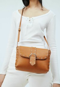 Borsa a tracolla in pelle color cuoio con borchie d'argento, chiusura a patta e dettaglio circolare in metallo. Tracolla regolabile e texture liscia.