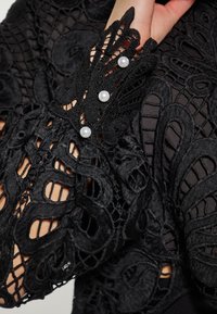 Blusa nera in pizzo con intricate fantasie floreali, maniche strutturate con tagli e tre bottoni in perla bianca sul polsino.
