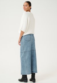 Weißer, flauschiger Pullover mit kurzen Ärmeln, kombiniert mit einem langen, hellblauen Jeansrock mit Taschen und Nahtdetails, getragen mit schwarzen Stiefeln.