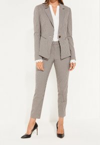 Completo gessato in grigio e bianco, giacca su misura con un solo bottone, pantaloni slim-fit coordinati e décolleté nere a punta.