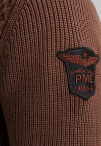 Bruin gebreid sweater met ribbeltextuur en een zwarte patch met rode borduursel, waarop vleugels en de tekst "PME 1649-A" staan afgebeeld.