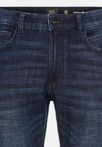 Jean en denim bleu foncé avec une coupe régulière, featuring une fermeture à boutons, des passants de ceinture et des coutures contrastantes. Texture lisse avec un léger effet délavé.
