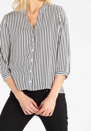 Frau trägt ein schwarz-weiß gestreiftes Hemd mit Knöpfen, drei Viertel Ärmel und schwarze Hose, die Hände vor sich verschränkt.