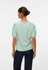 Blusa verde menta con maniche corte a sbuffo, intricati motivi a rombo con tagli, e scollatura rotonda. Indossata con jeans scuri.
