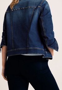 Giubbotto di jeans blu scuro con dettagli in borchie sulle spalle, caratterizzato da una vestibilità rilassata e maniche arrotolate, realizzato con cuciture rifinite.