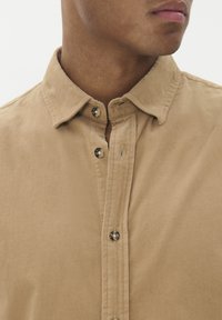 Smėlinis kordūro mėlynas marškiniai su užsegimu priekyje, smaigalio formos apykakle ir dviem matomais mygtukais. Tekstūruota paviršius su briaunuota struktūra.