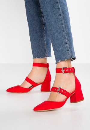 Rode suède hoge hakken met een spitse neus, voorzien van enkelbandjes en vierkante hakken. De schoenen hebben een gladde textuur en minimale detailing.