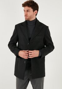 Tumši pelēkā vilnas maisījuma mētelis ar asu atlocītu apkakli, vienu pogu aizdari un priekšējām kabatām. Valkāts virs pieguļošas turtleneckreklu. Pielāgota piegriezums.