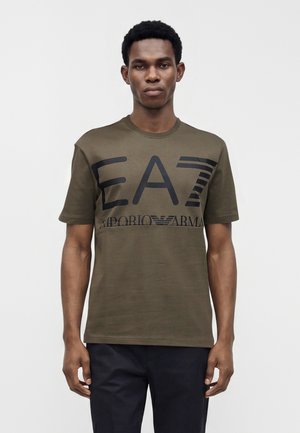 Homme à la peau foncée avec des cheveux courts et bouclés, portant un t-shirt vert olive EA7 Emporio Armani et un pantalon foncé, debout devant un fond uni.
