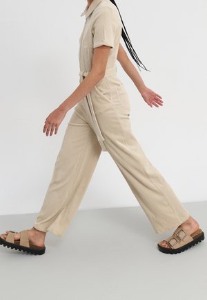 Beżowy sztruksowy kombinezon z krótkimi rękawami, kołnierzem i paskiem w talii. Modelka ma na sobie brązowe sandały z grubą czarną podeszwą.