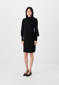 Juoda raukšlėta megzta suknelė su aukštu kaklu ir pūkuotomis ilgomis rankovėmis, iki kelių ilgio, derinama su juodais aukštakulniais slingback batais.