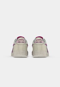 Sneakers featuring a beige synthetic upper with purple accents, textured rubber sole, and branded heel tab. Classic, retro design elements.