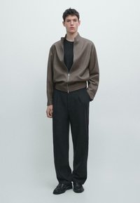 Braune Reißverschlussjacke über einem schwarzen Shirt, kombiniert mit locker sitzenden schwarzen Hosen und schwarzen Schuhen. Schlichtes Design, strukturierter Stoff.