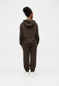 Γυναίκα που στέκεται γυρισμένη από πίσω, φορώντας σκούρο καφέ φούτερ με κουκούλα, αντίστοιχο παντελόνι φόρμας και άσπρα αθλητικά παπούτσια, μπροστά σε απλό λευκό φόντο.