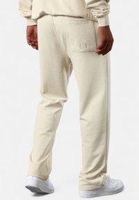 Beige Jogginghose aus strukturiertem Stoff, mit einer Gesäßtasche und dezenten Stickdetails sowie elastischen Bündchen. Getragen mit weißen Turnschuhen.