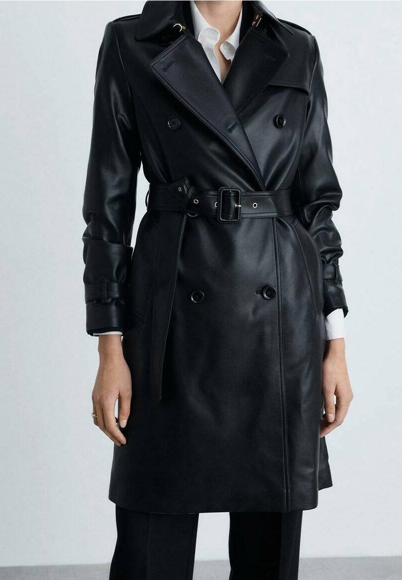 Personne portant un trench-coat en cuir noir à double boutonnage avec une ceinture, les poignets de chemise blanche visibles, et un pantalon noir visible sous le manteau.