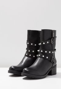 Botas negras hasta el tobillo hechas de material sintético texturizado, con dos diseños de correas con tachuelas de plata, cremalleras laterales y tacones cuadr ados.