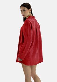 Rode textuursjerpjurk met lange mouwen, een kraag en een losse pasvorm, voorzien van een plooi aan de achterkant voor bewegingsvrijheid.