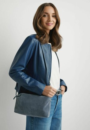 Kobieta w niebieskiej skórzanej kurtce i dżinsach trzyma szarą zamszową torbę na ramię jedną dłonią na jednolitym białym tle.