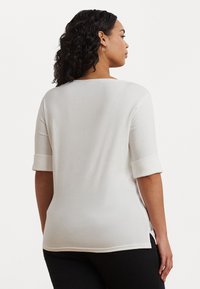 Λευκή μπλούζα κοντομάνικη με λεπτομέρειες σε ρολό, στρογγυλή λαιμόκοψη και πλαϊνές σχισμές, φτιαγμένη από ένα μαλακό ύφασμα με ελαφριά ελαστικότητα.