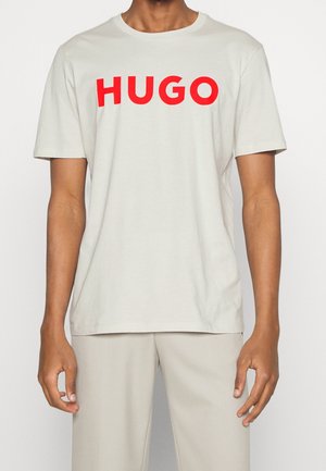 Homme portant un t-shirt beige uni avec un grand texte rouge "HUGO" sur la poitrine et un pantalon beige assorti, debout les bras détendus.