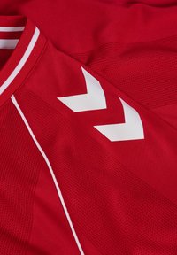 Czerwona sportowa koszulka z białym prążkowanym paskiem na kołnierzu, białym obszyciem i dwoma białymi kątowymi wzorami typu chevron na ramieniu.
