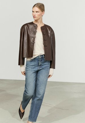 Woman wearing a dark brown leather cropped jacket with toggle buttons, light beige top, blue jeans, and pointed brown shoes walking indoors.