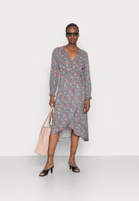Vestido com padrão floral, mangas longas, decote em V e barra assimétrica em preto, rosa e azul. Modelo segura uma bolsa de mão rosa claro.