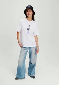 Balta pārsteidzoša tees ar drukātu dizainu, kombinēta ar gaiši ziliem plašiem džinsiem un melnām punktiņotām kurpēm. Iekļauta melna bļodiņa cepure.