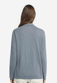 Grå långärmad skjorta med krage, tillverkad av mjukt stickat material. Har en rak fåll och minimal textur, utan synliga mönster eller detaljer.