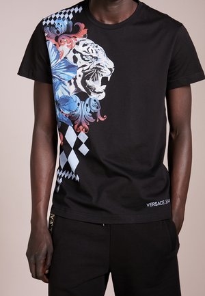 T-shirt noir avec un tigre blanc rugissant, motifs floraux bleus et roses et à carreaux sur la poitrine gauche, porté par une personne à la peau foncée.