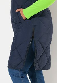 Granatowa pikowana kamizelka z bocznym zamkiem błyskawicznym, wyposażona w kieszeń po lewej stronie i wewnętrzną wyściółkę w kontrastującym kolorze. Rękawy w jaskrawym neonowym zielonym kolorze.