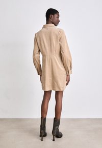 Kordhemdkleid in hellem Beige mit Kragen, langen Ärmeln und A-Linien-Silhouette. Kombiniert mit schwarzen, Absatzstiefeletten.