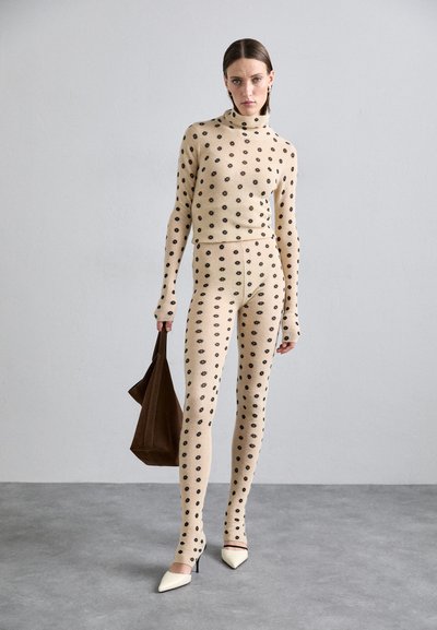 Completo in maglia a due pezzi beige con motivi floreali neri; presenta un top a collo alto e pantaloni aderenti, abbinati a una borsa marrone.