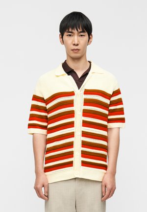 Jeune homme portant une chemise tricotée à manches courtes à rayures crème, orange, rouge et marron, boutonnée, par-dessus un polo foncé, debout devant un fond blanc.