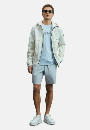 Veste blanche légère avec fermeture éclair à l'avant, superposée à un t-shirt bleu clair, associée à un short gris clair et des baskets blanches.