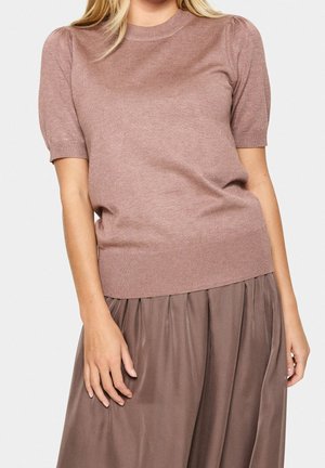 Mujer vistiendo una blusa de punto corta en color malva combinada con una falda plisada malva a juego, de pie con una postura relajada frente a un fondo blanco.