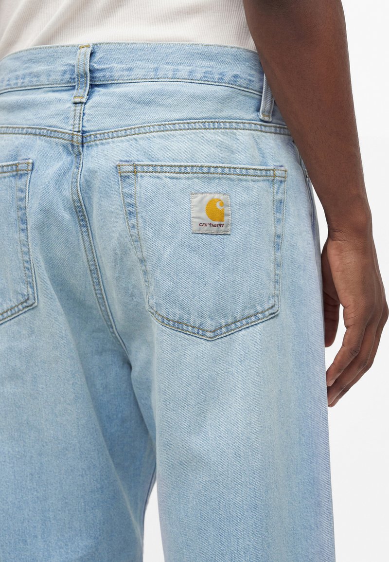 Light blue denim jeans with a visible Carhartt logo patch on the back right pocket, worn by a person with dark skin and white top.