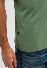 Groene katoenen t-shirt met ronde hals, korte mouwen en een zijtag detail. De stof is soepel met een lichte stretch. Denim jeans zichtbaar eronder.