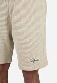 Beige teksturerede shorts lavet af letvægtsstof, med en afslappet pasform og et lille sort logo på nederste venstre side.